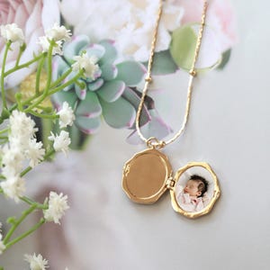 May include: A gold locket necklace with a small photograph inside. The locket is open, displaying a baby's picture. The necklace has a delicate chain with small gold beads. The background features white flowers and greenery.