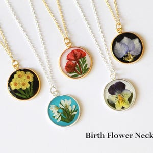 May include: A collection of round pendant necklaces featuring pressed flowers encased in resin. The pendants are on gold and silver chains. The flowers are yellow, red, white, blue, and purple. The text "Birth Flower Necklace" is at the bottom.