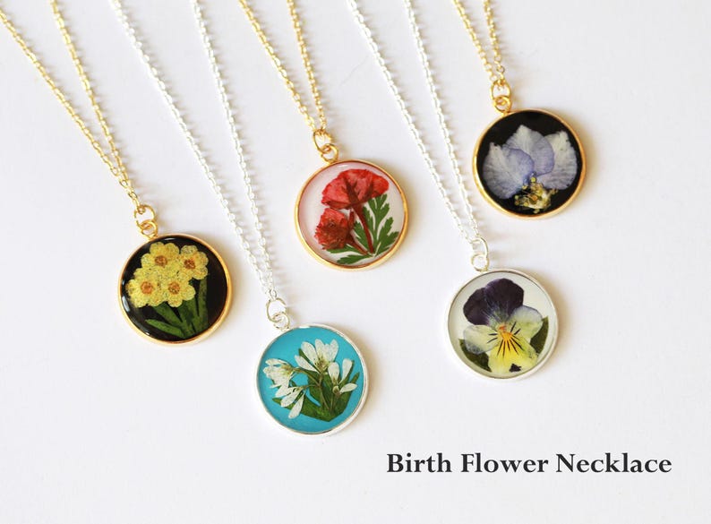May include: A collection of round pendant necklaces featuring pressed flowers encased in resin. The pendants are on gold and silver chains. The flowers are yellow, red, white, blue, and purple. The text "Birth Flower Necklace" is at the bottom.