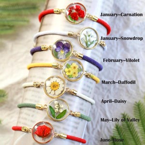 May include: A collection of bracelets featuring pressed flowers encased in clear resin. Each bracelet has a different flower, including carnation, snowdrop, violet, daffodil, daisy, lily of the valley, and rose. The bracelets are labeled with the month and flower name.