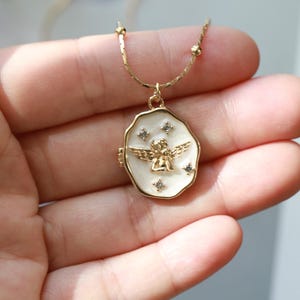 May include: Gold-coloured necklace with a pendant featuring a white enamel background and a gold angel figure. The pendant is adorned with small, sparkling crystals. The necklace is held in a hand.