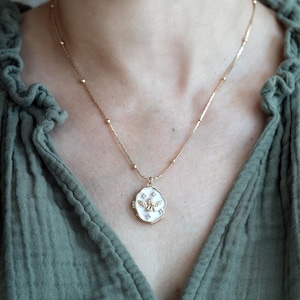 May include: A gold-coloured necklace with a pendant featuring a white background and a gold-coloured angel design. The pendant is surrounded by small, white star-shaped accents. The necklace is worn against a green top.