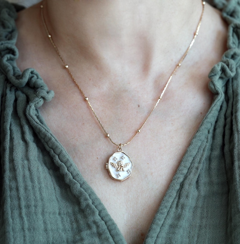 May include: A gold-coloured necklace with a pendant featuring a white background and a gold-coloured angel design. The pendant is surrounded by small, white star-shaped accents. The necklace is worn against a green top.
