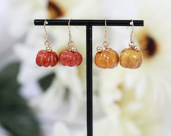May include: Four pairs of pumpkin-shaped earrings, two pairs are orange and two pairs are yellow. The earrings are hanging from a black stand.