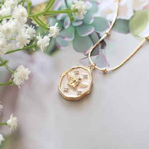 May include: Gold-coloured necklace with a pendant featuring a winged figure and small crystals. The pendant has a white enamel background and a gold-coloured border. Delicate chain with small gold beads.