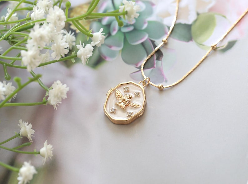 May include: Gold-coloured necklace with a pendant featuring a winged figure and small crystals. The pendant has a white enamel background and a gold-coloured border. Delicate chain with small gold beads.