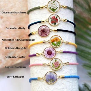 May include: A collection of seven bracelets featuring pressed flowers encased in clear resin, each with a gold-colored bezel. The bracelets have colorful cords in shades of green, blue, black, yellow, purple, pink, and blue. Text on the image indicates the flower and month.