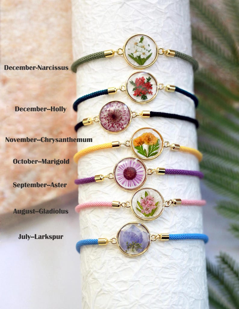 May include: A collection of seven bracelets featuring pressed flowers encased in clear resin, each with a gold-colored bezel. The bracelets have colorful cords in shades of green, blue, black, yellow, purple, pink, and blue. Text on the image indicates the flower and month.