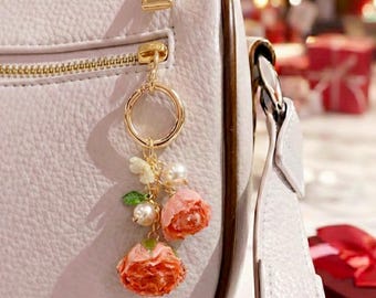 May include: A light gray handbag with a gold zipper and a decorative floral keychain. The keychain features two orange-pink roses, pearls, and a small butterfly charm. The bag has a white strap.