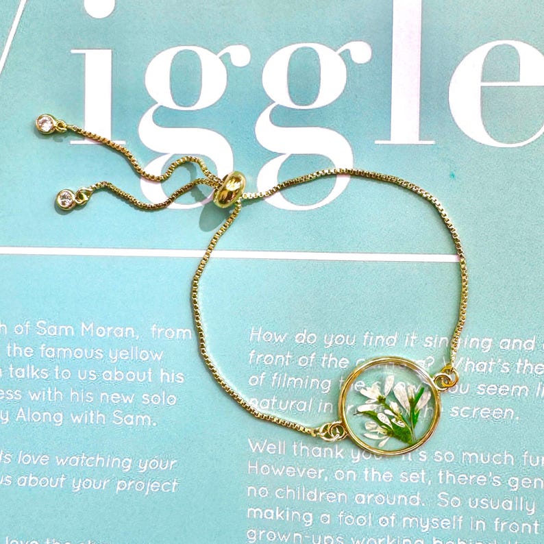 May include: Gold-toned adjustable bracelet featuring a clear, round charm with delicate white flowers preserved inside. The bracelet has a delicate chain and small, sparkling accents near the clasp.