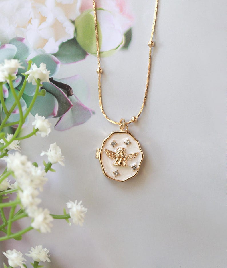May include: A gold-colored necklace with a pendant featuring an angel design. The pendant is round with a white enamel background and gold trim. The angel is gold-colored with outstretched wings. The necklace chain has small gold beads and a delicate design. The background includes white flowers and a soft, neutral color palette.