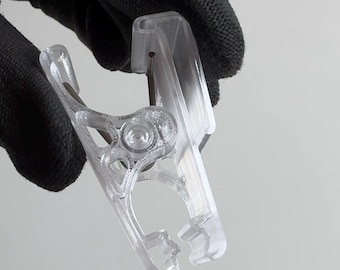 May include: A clear plastic spring-loaded clamp featuring a hinged design with a metal coil spring at the pivot point. The clamp has a textured grip area and a specialized notched jaw structure at the tip for securing items in place.