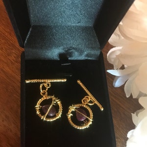 May include: Gold-coloured cufflinks with a circular design, each featuring a small, dark red gemstone. The cufflinks are presented in a black velvet box. The design is intricate and elegant, suitable for formal wear or as a gift.