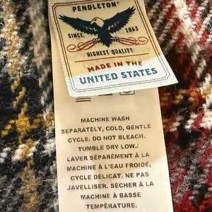May include: A Pendleton brand fabric label attached to a red, black, and white plaid textile. The label features an eagle graphic, the text PENDLETON SINCE 1863 HIGHEST QUALITY MADE IN THE UNITED STATES, and care instructions in English and French.