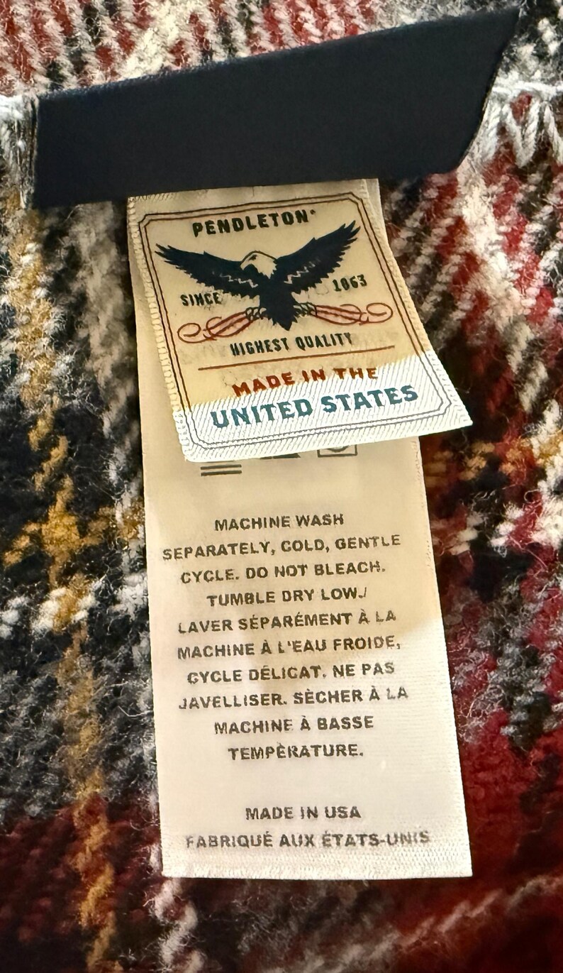 May include: A Pendleton brand fabric label attached to a red, black, and white plaid textile. The label features an eagle graphic, the text PENDLETON SINCE 1863 HIGHEST QUALITY MADE IN THE UNITED STATES, and care instructions in English and French.