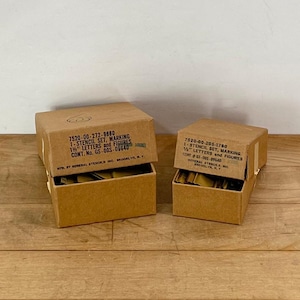 May include: Two open cardboard boxes containing stencil sets. The boxes are labeled with text including "1-STENCIL SET, MARKING 1 1/2" LETTERS and FIGURES". The boxes are on a wooden surface.