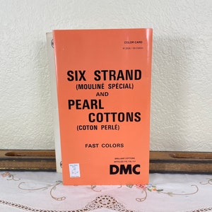 May include: A bright orange DMC color card for six strand mouliné spécial and pearl cottons. The front cover features black text reading Six Strand Mouliné Spécial and Pearl Cottons Coton Perlé Fast Colors, with the DMC logo at the bottom right.