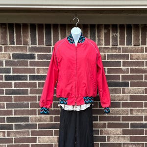 May include: A red bomber jacket with a zipper closure, white piping, and patterned trim in blue, black, and turquoise. The jacket is displayed on a white mannequin head, paired with a floral shirt and black pants.