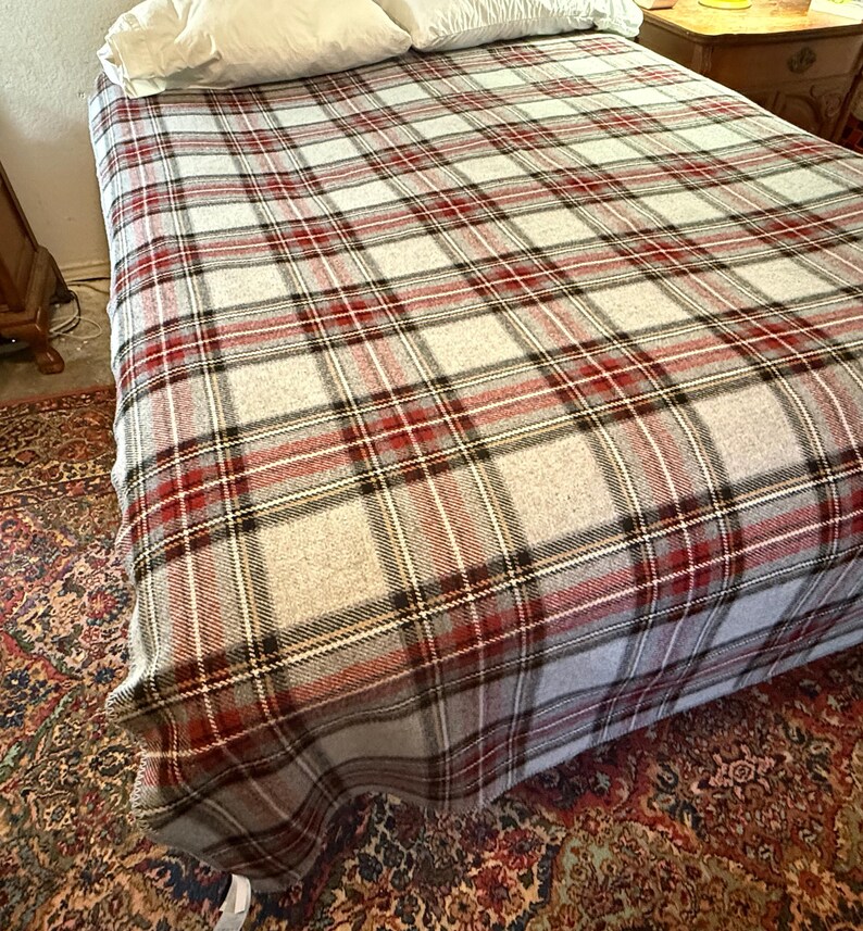 May include: A bed blanket featuring a classic plaid pattern in shades of light gray, deep red, and black. The woven fabric displays a repeating grid design that covers the entire surface of the bed, providing a textured appearance across the bedding.