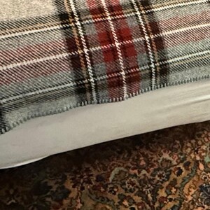 May include: A woven blanket featuring a plaid pattern in shades of red, black, white, and light gray. The fabric has a textured appearance with visible intersecting lines forming a classic grid design across the entire surface of the textile.
