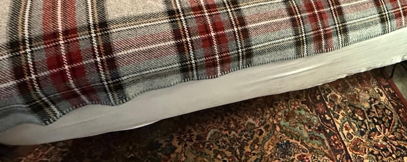 May include: A woven blanket featuring a plaid pattern in shades of red, black, white, and light gray. The fabric has a textured appearance with visible intersecting lines forming a classic grid design across the entire surface of the textile.