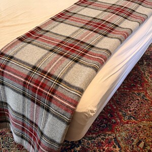 May include: A rectangular throw blanket featuring a classic plaid pattern in shades of light gray, deep red, black, and thin white lines. The fabric has a visible woven texture and is draped across the surface of a bed with white bedding.