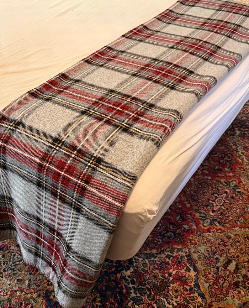 May include: A rectangular throw blanket featuring a classic plaid pattern in shades of light gray, deep red, black, and thin white lines. The fabric has a visible woven texture and is draped across the surface of a bed with white bedding.