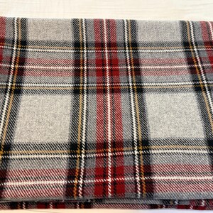 May include: A folded rectangular blanket featuring a classic plaid pattern. The design consists of intersecting horizontal and vertical stripes in shades of light gray, deep red, black, and thin white and yellow accent lines throughout the fabric.