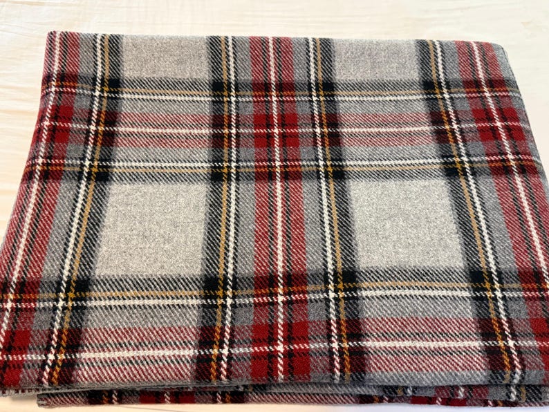 May include: A folded rectangular blanket featuring a classic plaid pattern. The design consists of intersecting horizontal and vertical stripes in shades of light gray, deep red, black, and thin white and yellow accent lines throughout the fabric.