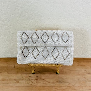 May include: A white beaded clutch with a diamond pattern, resting on a gold-colored stand. The clutch is rectangular with a fold-over flap. The diamonds are outlined in a darker color.
