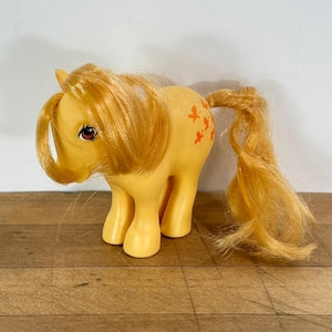 May include: A vintage yellow toy pony with a long, flowing orange mane and tail. The pony has orange butterfly markings on its side and brown eyes. The toy is standing on a wooden surface.
