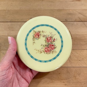 May include: A round, pale yellow compact with a floral design. The compact features a teal blue border and a cluster of pink roses with green leaves. The compact is held in a hand.
