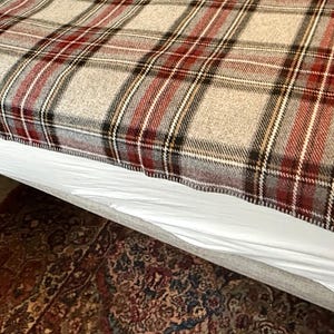 May include: A woven blanket featuring a classic plaid pattern in shades of beige, deep red, black, and white. The fabric displays a textured, soft appearance with a consistent grid design that covers the entire surface of the rectangular textile.