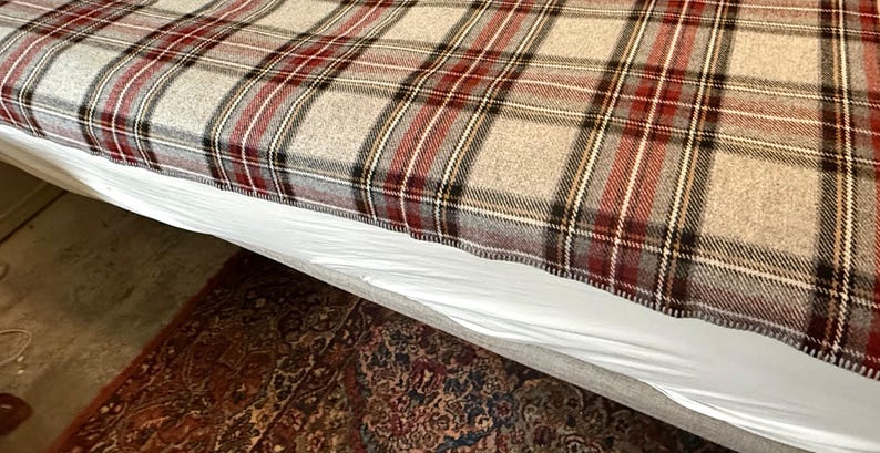 May include: A woven blanket featuring a classic plaid pattern in shades of beige, deep red, black, and white. The fabric displays a textured, soft appearance with a consistent grid design that covers the entire surface of the rectangular textile.