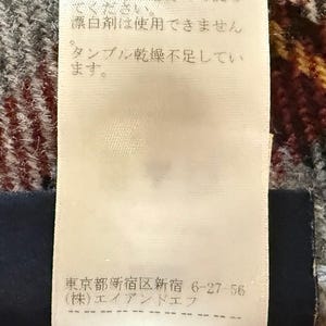 May include: A white rectangular garment care label with black Japanese text and laundry symbols. The label includes washing instructions, a prohibition on bleach, and a company address in Shinjuku, Tokyo, printed on a textured fabric surface.