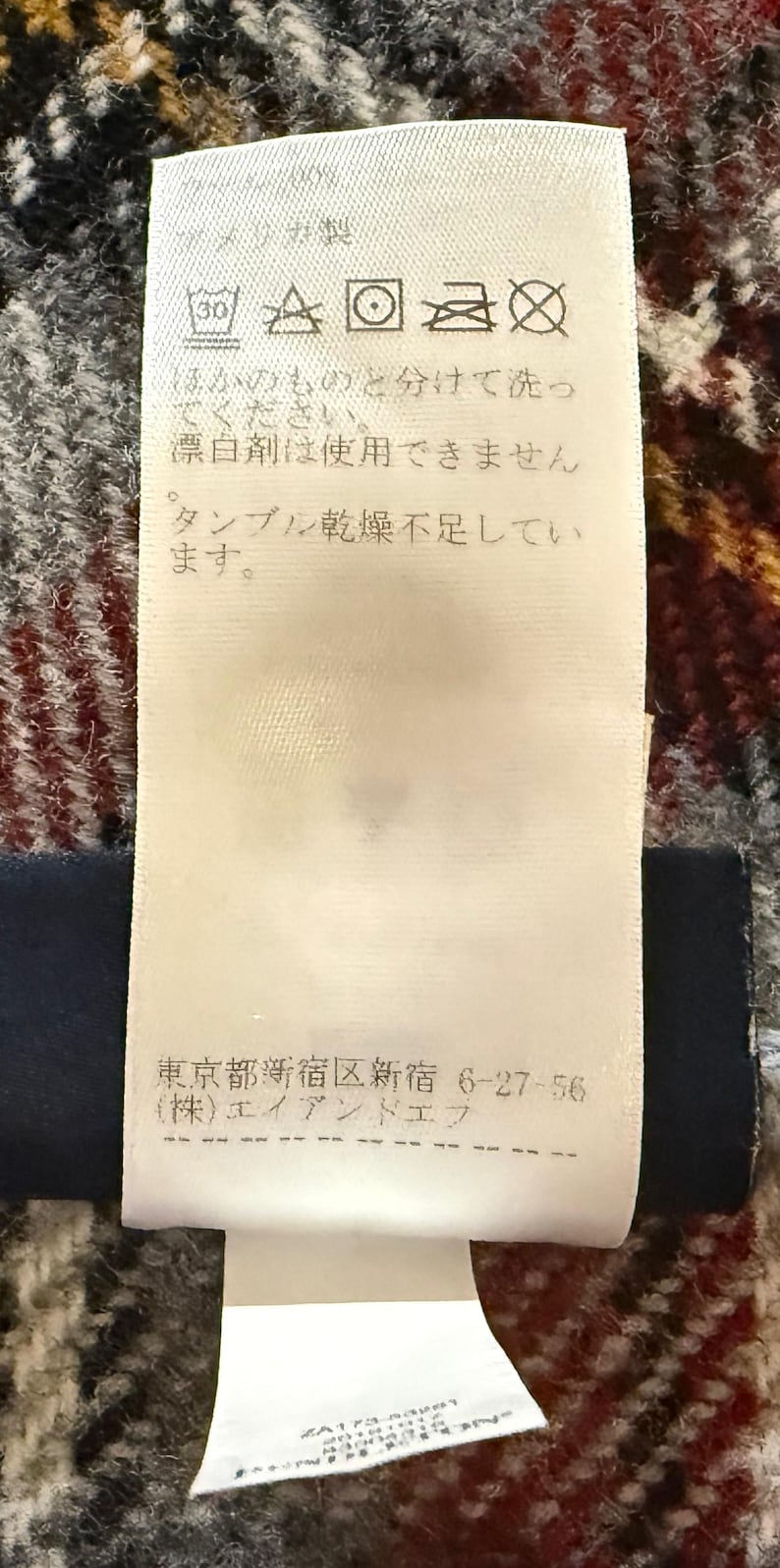 May include: A white rectangular garment care label with black Japanese text and laundry symbols. The label includes washing instructions, a prohibition on bleach, and a company address in Shinjuku, Tokyo, printed on a textured fabric surface.