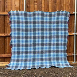 May include: A blue, gray, and black plaid blanket with red fringe. The blanket is hanging on a wooden fence. The plaid pattern is made up of squares and rectangles of varying sizes.