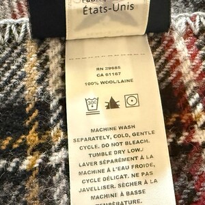 May include: A Pendleton brand clothing label attached to a plaid wool fabric. The cream-colored tag features black text including "Depuis 1863," "100% Wool," and "Made in USA," along with care instructions for machine washing, no bleaching, and tumble drying.