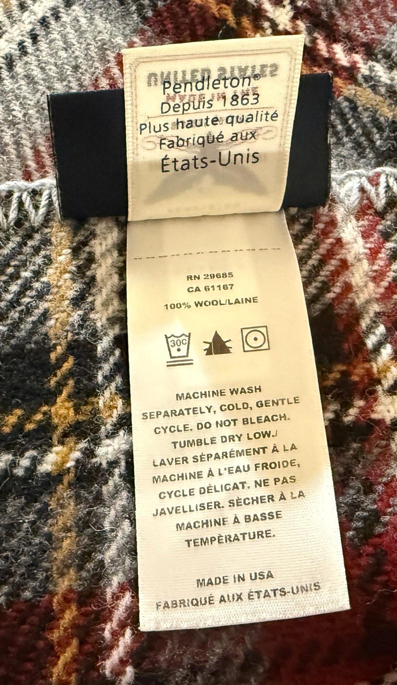 May include: A Pendleton brand clothing label attached to a plaid wool fabric. The cream-colored tag features black text including "Depuis 1863," "100% Wool," and "Made in USA," along with care instructions for machine washing, no bleaching, and tumble drying.