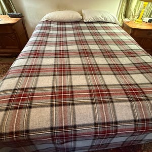 May include: A bed featuring a light gray blanket with a prominent plaid pattern in shades of dark red, black, and white. Two plain white rectangular pillows are positioned at the head of the bed, resting against the top edge of the plaid blanket.