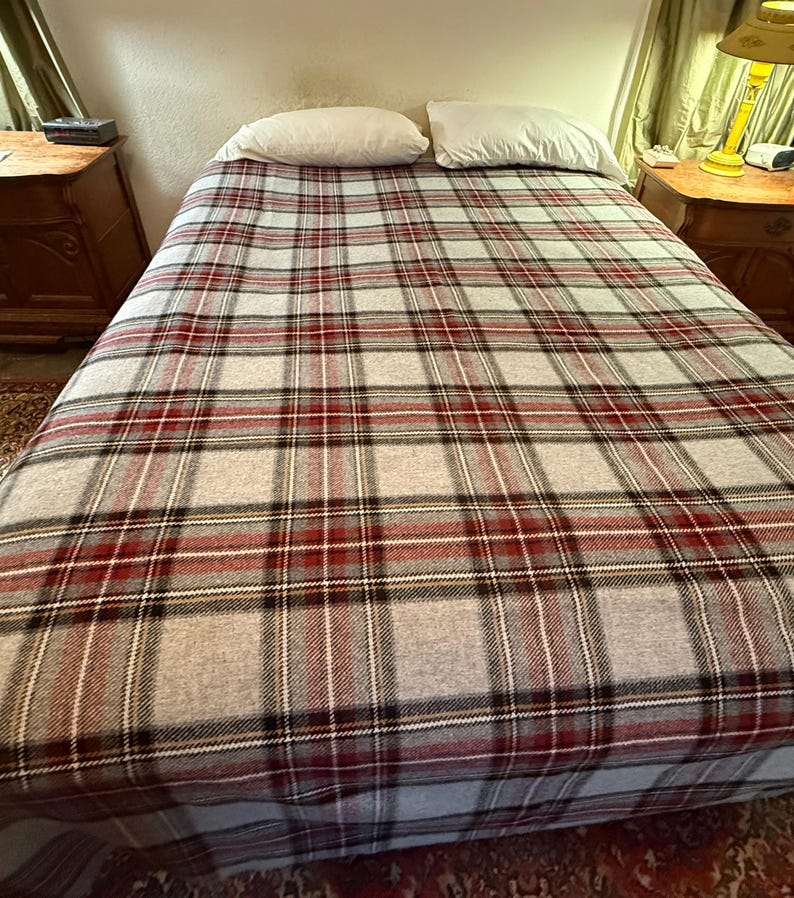 May include: A bed featuring a light gray blanket with a prominent plaid pattern in shades of dark red, black, and white. Two plain white rectangular pillows are positioned at the head of the bed, resting against the top edge of the plaid blanket.