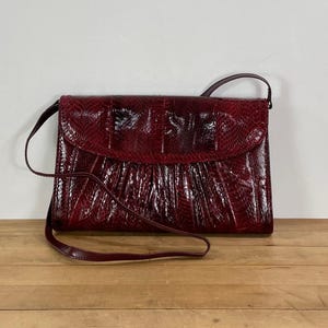 May include: A burgundy-colored handbag with a flap closure and a long shoulder strap. The bag has a textured, reptile-like pattern. The strap is thin and made of the same material as the bag. The bag is rectangular in shape.