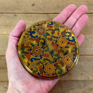 May include: A round, gold-toned compact with a floral and geometric design. The compact features orange, brown, blue, and green accents. The design includes stylized flowers, triangles, and curved lines. The compact is held in a hand.