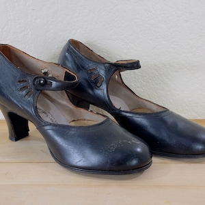 May include: A pair of vintage black leather shoes with a Mary Jane style. The shoes have a strap across the top of the foot and a small heel. The sides of the shoes have decorative cutouts.