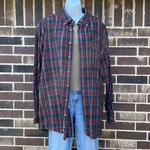 May include: A button-down plaid shirt in burgundy, navy, and green, displayed on a mannequin. The shirt is paired with a tan undershirt and light blue jeans. The shirt has long sleeves and a classic collar.