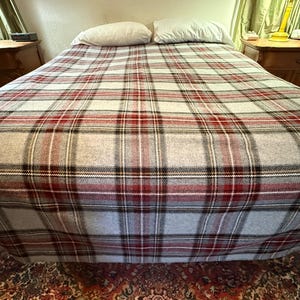May include: A bed covered with a plaid blanket featuring a pattern of intersecting red, black, and white lines on a light gray background. Two plain white pillows are positioned at the head of the bed, resting against a light-colored wall.