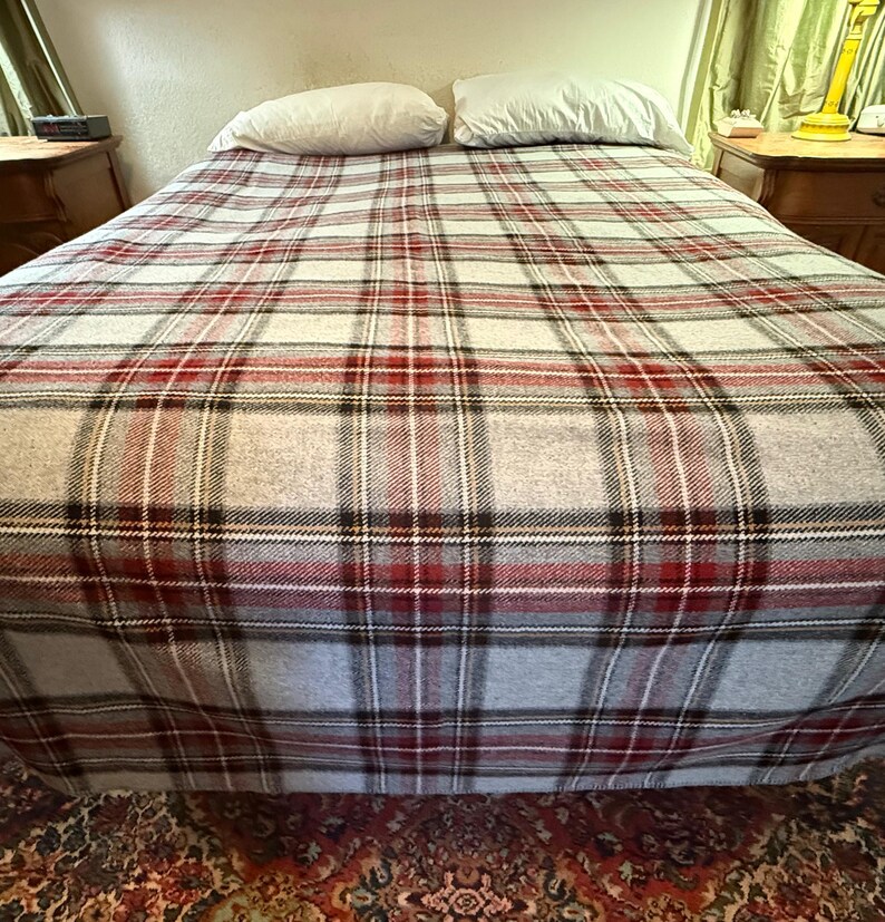 May include: A bed covered with a plaid blanket featuring a pattern of intersecting red, black, and white lines on a light gray background. Two plain white pillows are positioned at the head of the bed, resting against a light-colored wall.