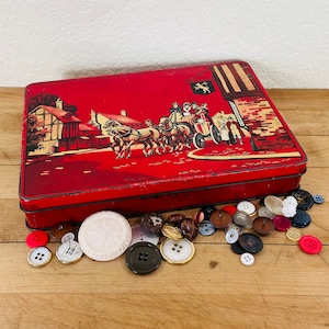 May include: A red rectangular tin with a vintage illustration of a horse-drawn carriage and buildings. Assorted buttons in various colors and sizes are scattered around the tin, including red, black, and white buttons.