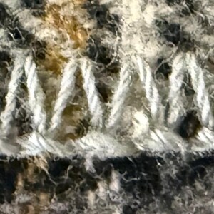 May include: A close-up view of a fabric edge finished with a white blanket stitch. The textile features a plaid pattern with interwoven threads in shades of gray, black, and red, showing a textured, fuzzy surface characteristic of wool or flannel.