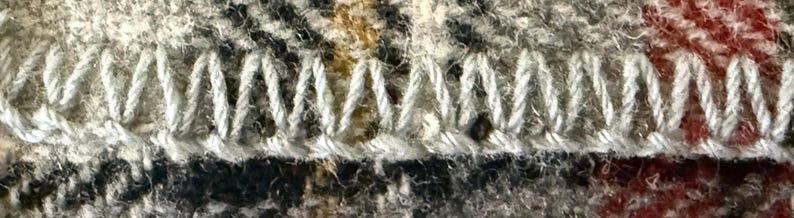 May include: A close-up view of a fabric edge finished with a white blanket stitch. The textile features a plaid pattern with interwoven threads in shades of gray, black, and red, showing a textured, fuzzy surface characteristic of wool or flannel.